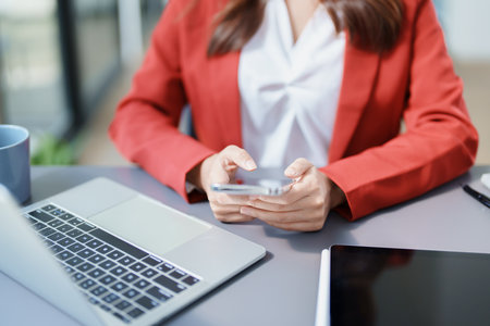 Portrait of a young Asian woman showing a smiling face as she uses her phone, computer and financial documents on her desk in the early morning hours.の写真素材