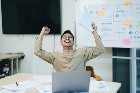 Portrait of a male business owner showing a happy smiling face as he has successfully invested his business using computers and financial budget documents at work.の写真素材