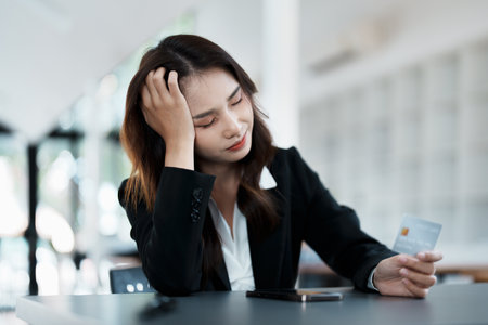 Portrait of young Asian woman holding credit card with smart phone mobile and showing worried expression on credit card debtの写真素材