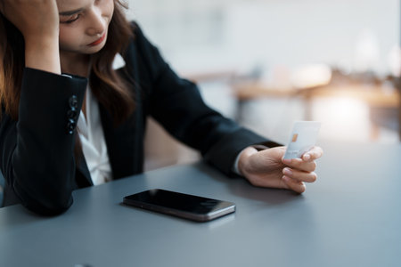Portrait of young Asian woman holding credit card with smart phone mobile and showing worried expression on credit card debtの写真素材
