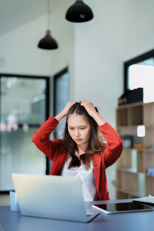 Portrait of a young Asian woman showing a serious face as she uses her phone, financial documents and computer laptop on her desk in the early morning hours.の写真素材