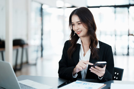 Portrait of a young Asian woman showing a smiling face as she uses her phone, computer and financial documents on her desk in the early morning hours.の写真素材