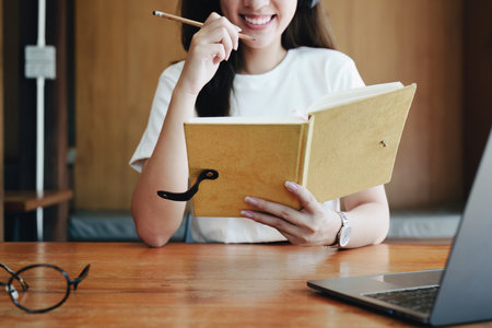 Portrait of a teenage Asian woman using a computer, wearing headphones and using a notebook to study online via video conferencing on a wooden desk in library.の写真素材
