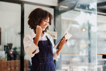 Startup successful small Beautiful American African owner sme with tablet in coffee shop restaurant, hands up in winner is gesture, Happy to be successful celebrating achievement success.の写真素材