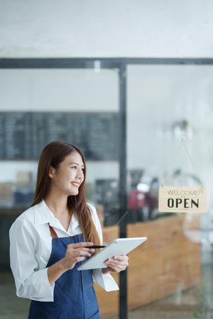 Portrait of a beautiful Asian woman running a small business holding a computer tablet showing a smiling face, a coffee shop owner opening a shop to welcome customers in the new morning, SME concepts.の写真素材