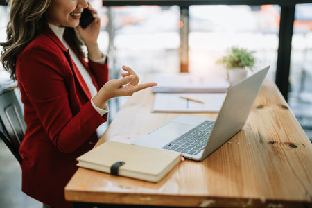 Portrait of a young Asian woman showing a smiling face as she uses her phone, computer and financial documents on her desk in the early morning hours.の写真素材