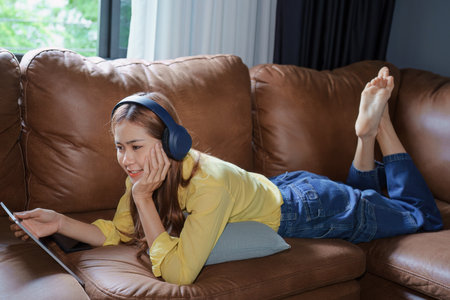 Portrait of asian woman using tablet and headphones relaxing on sofa at home.の写真素材