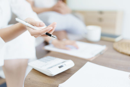 Portrait of young Asian female agent offering life insurance to client using document, calculator and tablet in product presentation.の写真素材