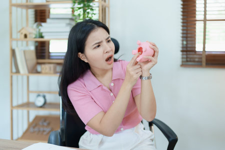 Portrait of an Asian female businesswoman showing a serious and anxious expression in an SME business , but no money in the pink pig piggy bank , saving plans.の写真素材