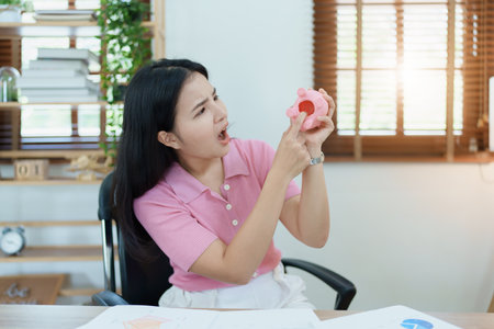 Portrait of an Asian female businesswoman showing a serious and anxious expression in an SME business , but no money in the pink pig piggy bank , saving plans.の写真素材