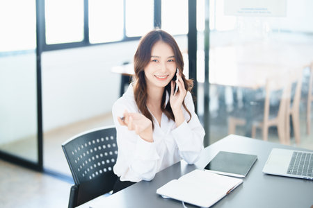 Portrait of a young Asian man showing a smiling face as she uses his phone, computer and financial documents on her desk in the early morning hours.の写真素材