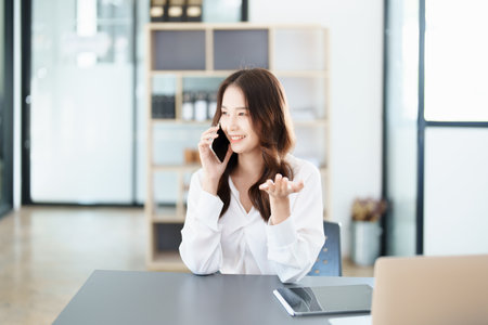 Portrait of a young Asian man showing a smiling face as she uses his phone, computer and financial documents on her desk in the early morning hours.の写真素材
