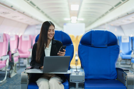 Asian business woman passenger sitting on business class luxury plane while working using smart phone mobile talking and use laptop computer with simulated space using on board wireless connectionの写真素材