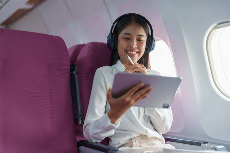 Young Asian attractive business woman passenger sitting on business class luxury plane while wearing wireless headphone for relax and working with tablet during the flight.の写真素材