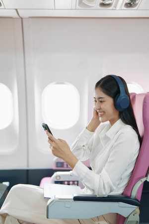 Young Asian attractive business woman passenger sitting on business class luxury plane while wearing wireless headphone and using smart phone mobile for relax during the flight.の写真素材