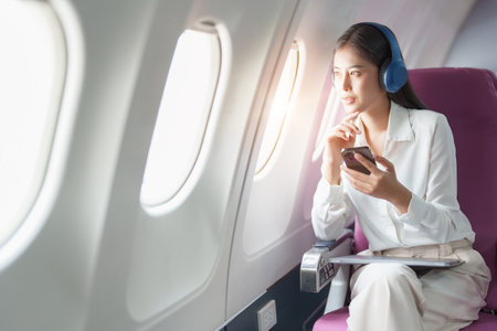 Young Asian attractive business woman passenger sitting on business class luxury plane while wearing wireless headphone and using smart phone mobile for relax during the flight.の写真素材