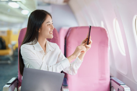 Young Asian attractive business woman passenger sitting on business class luxury plane while using smart phone mobile to take a photo and use computer laptop during the flight.の写真素材