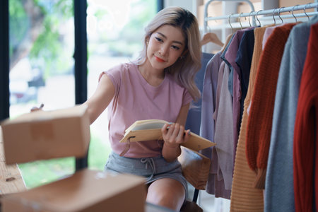 Portrait Of Asian Female Owner Of Fashion Store Checking Stock In Clothing Store With using notebook successful happy smile at small business, sme or ecommerce concepts.の写真素材
