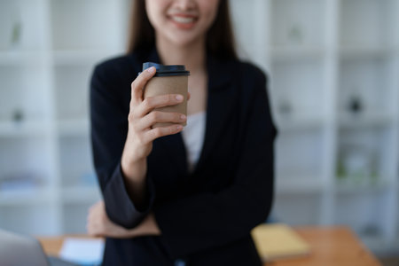 Young minded successful employee business woman in black suit work hold paper cup coffee to go stand at desk with laptop computerの写真素材