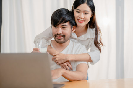 Happy young loving family couple looking at laptop computer screen, reading ecipes. Wife showing embrace to husband to pretty joyful husband, sitting at table in kitchen at home in weekendの写真素材