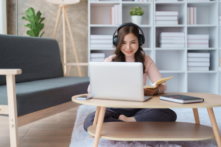 Portrait of a teenage Asian woman using a laptop computer, wearing headphones and using a notebook...