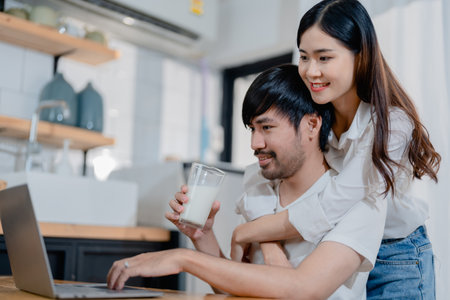 Happy young loving family couple looking at laptop computer screen, reading ecipes. Wife showing embrace to husband to pretty joyful husband, sitting at table in kitchen at home in weekendの写真素材