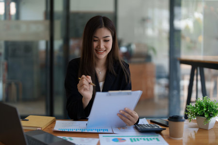 Young asian businesswoman manager, lawyer or company employee holding accounting bookkeeping documents checking financial data or marketing report working in office with laptop. Paperwork management.の写真素材