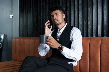 Portrait of a nice and discreet Asian man sitting on a sofa using a smartphone and drinking coffee.