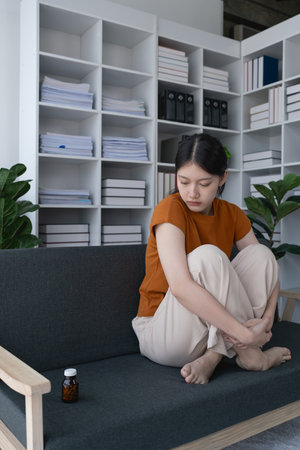 The woman, sitting on the sofa at home, stared at the medicine bottle, a symbol of her battle with depression.の写真素材