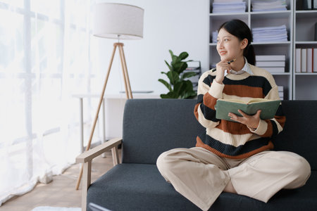 A woman sits on a sofa, using her notebook and tablet computer simultaneously to study online,...