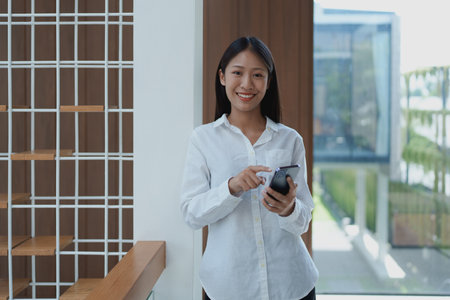 A young woman in a stylish office smiles while using her smartphone. The image highlights modern workspace and technology in professional life with natural lighting.の写真素材