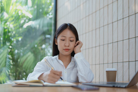 A focused woman writes in her notebook at a cozy wooden table, enjoying a coffee while surrounded by...
