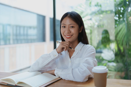 A cheerful young woman enjoys a coffee while contemplating ideas at a wooden table, surrounded by plants and natural light, creating a peaceful and inspiring workspace ambiance.の写真素材
