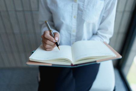 Individual writing in notebook with pen in modern office environment, focused on creative and productive work.の写真素材