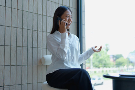 Professional woman communicating on smartphone with bright smile in modern office setting, enjoying conversation.の写真素材