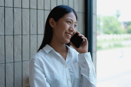 Young woman smiling while speaking on a smartphone against a modern interior wall with natural light...