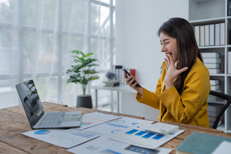 Happy young woman with smartphone at desk celebrating success in a modern office workspace filled with documents and plants.の写真素材