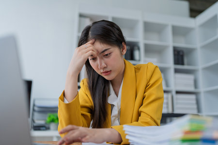 Stressed businesswoman in yellow blazer overwhelmed by work at a desk with papers and laptop in a modern office setting.の写真素材
