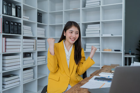 Excited young businesswoman celebrating success while checking smartphone at modern office desk with business reports.の写真素材