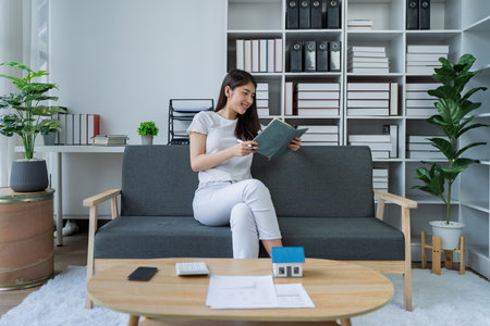 Young woman sitting on couch enjoying a book while planning finances in bright modern home office space.の写真素材