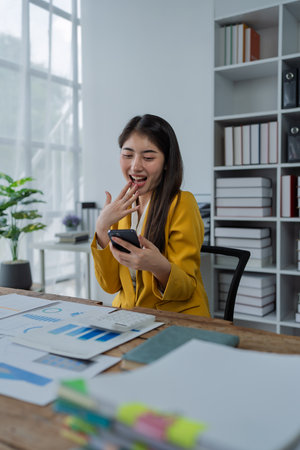 Happy young woman with smartphone at desk celebrating success in a modern office workspace filled with documents and plants.の写真素材