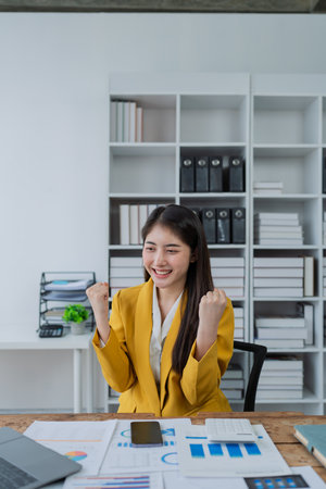 Excited young businesswoman celebrating success while checking smartphone at modern office desk with business reports.の写真素材
