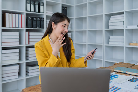 Happy young woman with smartphone at desk celebrating success in a modern office workspace filled with documents and plants.の写真素材