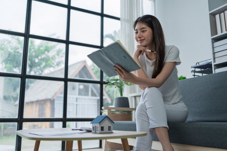 Young woman sitting on couch enjoying a book while planning finances in bright modern home office space.の写真素材