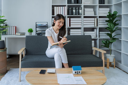 Young woman sitting on couch enjoying a book while planning finances in bright modern home office space.の写真素材