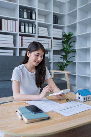 Young woman analyzing financial documents at home with calculator and model house on table in modern living room.の写真素材