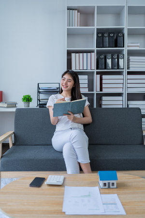 Young woman analyzing financial documents at home with calculator and model house on table in modern living room.の写真素材