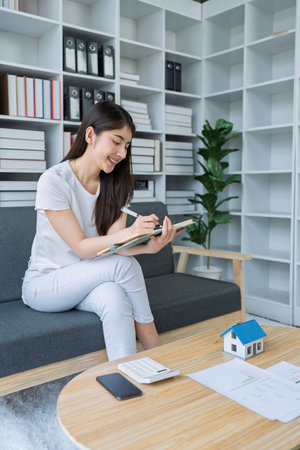 Young woman analyzing financial documents at home with calculator and model house on table in modern living room.の写真素材