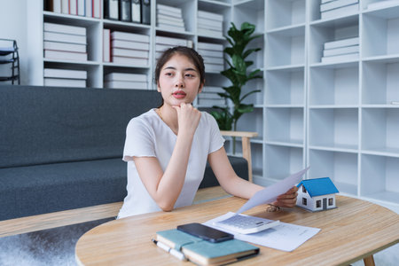 Young woman analyzing financial documents at home with calculator and model house on table in modern living room.の写真素材