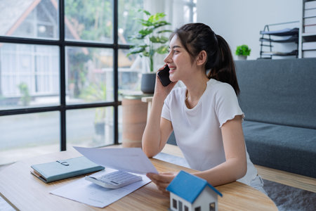 Young woman engaged in a phone conversation while working on home finances in a cozy modern living room.の写真素材